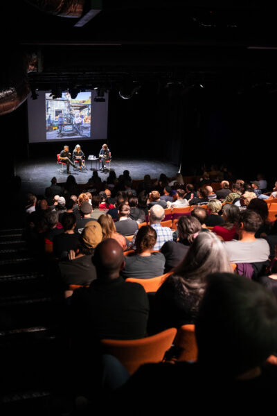 A group of people sit in an auditorium, listening to people on stage talking.