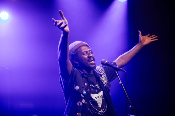A black person raises their hands in the arm - backlit by purple lights.