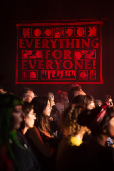 People sit in a crowd, behind them is a red and black banner than reads 'Everything for Everyone'.