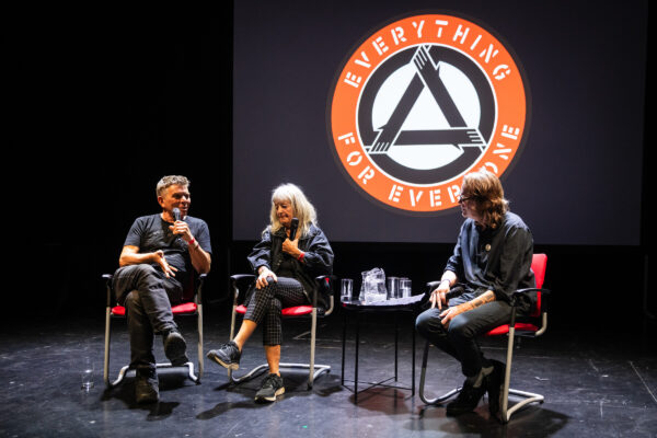 Three people talk on stage - behind them is a logo.