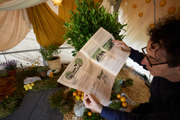 A man with dark hair and glasses reads a newspaper inside of the exhibition: a refugee tent with trees and plants.