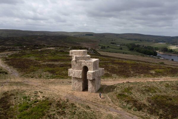 The artwork Tower by Steve Messam, in the landscape of Penistone Hill. The large sculpture is covered in sheep fleece, creating the shape of an arch with angular corners, set against a cloudy sky and the natural landscape.
