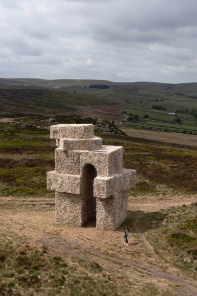 The artwork Tower by Steve Messam, in the landscape of Penistone Hill. The large sculpture is covered in sheep fleece, creating the shape of an arch with angular corners, set against a cloudy sky and the natural landscape.