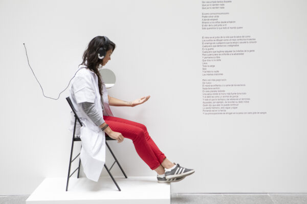 A woman with dark hair sits on a chair with headphones on, looking at her hand. She is wearing a white lab coat over a grey long-sleeved t-shirt and red trousers. There is text on the wall to the right of her and behind her, there is a hole in the wall.
