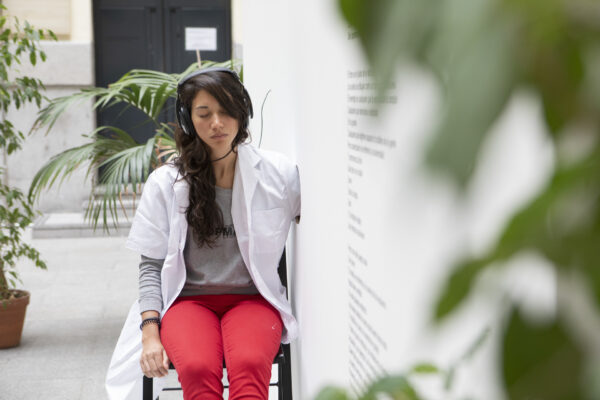 A woman with dark hair sits on a chair with headphones on, looking at her hand. She is wearing a white lab coat over a grey long-sleeved t-shirt and red trousers. She is listening with her eyes closed and her arm is through the hole in the wall - she is surrounded by plants.