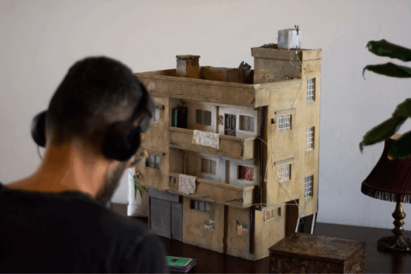A man with short hair bows his head as he listens to headphones. In focus in the background, there is a brown model of a building sat on a table beside a box and a short lamp.