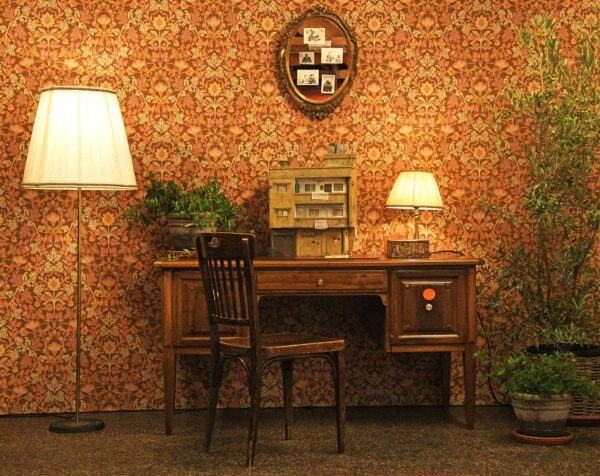 A desk and chair sit in the centre, with a model of a house on the desk. The are two lamps, some plants and the background is a vibrantly patterned orange wallpaper - on which is hung a oval picture frame containing sepia tone pictures.