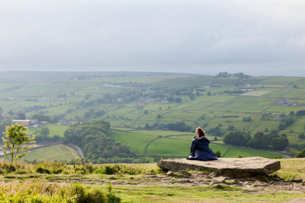 A view across the moors from Penistone Hill. A woman sits alone on a rock looking at the view.