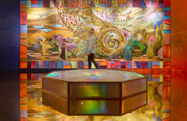 Zadie Xa's Turner Prize installation. A large octagonal piece, around knee height, is in front of us. Behind is a large colourful painting on the wall and a person wakling past looking at the artwork.