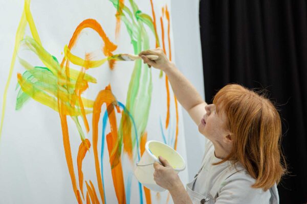 A red headed woman in a white overall paints patterns in green yellow and orange on a white canvas.