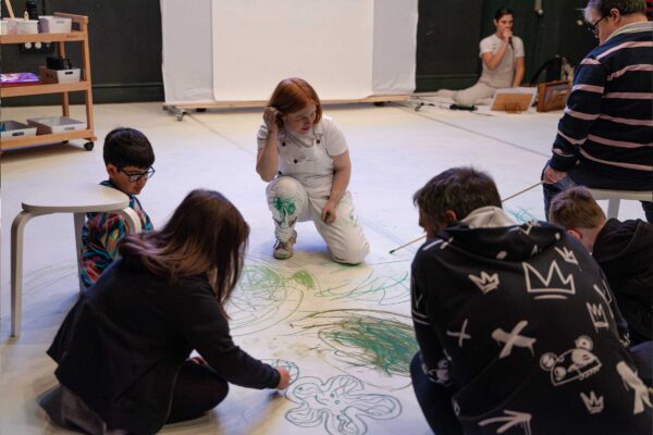 A group of people scrouch on a giant piece of paper, colouring it in with paint and pastels