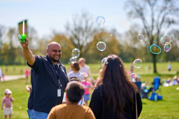 A amn smiling to a lady and young boy on a bright summers day in the park. The man is blowing bubbles.