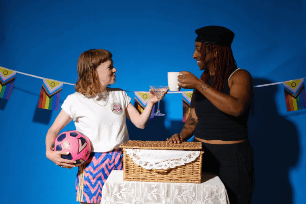 Two people pose for the camera - toasting with a glass and a mug. The person on the left is wearing a white t-shirt and shorts, and is holding a pink football. The person on the right is wearing a black hat and outfit. Between them, a picnic basket sits on a table and there is pride flag bunting behind.