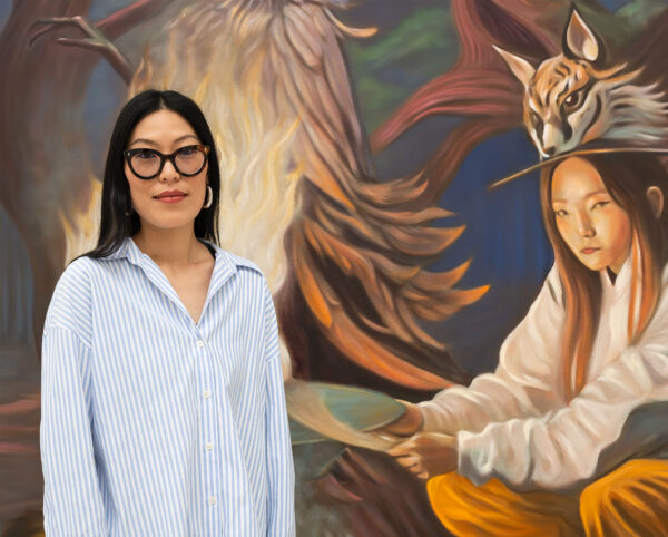 Artist Zadie Xa looks towards us. She is standing in front of a painting that shows a woman with wildlife including a large cat.