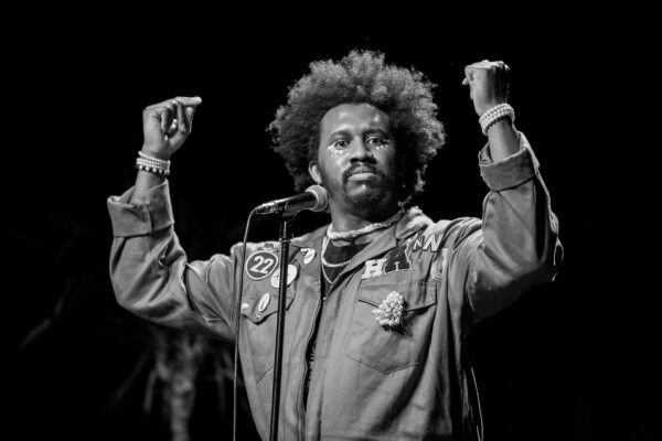 A black and white photo of a black man with combed out curls standing in front of a microphone with his hands in the air, fingers pointing to himself.