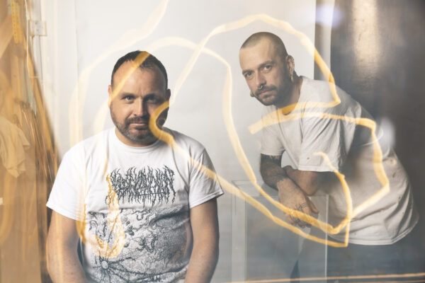 Two white men pose for the camera - they're wearing white t-shirts and are looking straight ahead. There is a soft focus on the camera and a warm golden neon blur is on the front of the image.