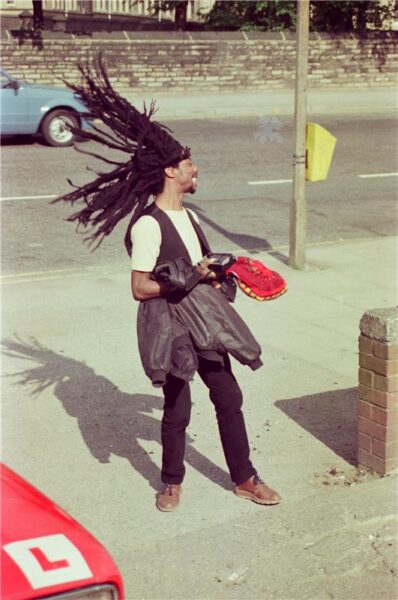 A man with long dreads flicks his hair, while standing on the pavement