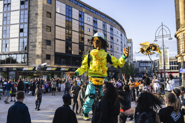 Zee, in a yellow and blue tracksuit surrounded by audience members who are stood in a circle.