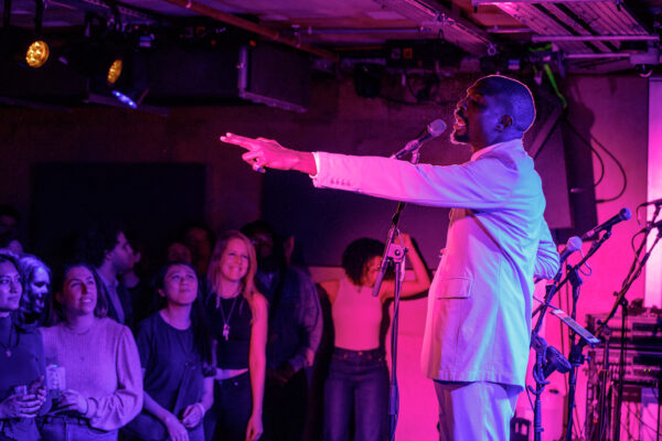 Man white suite, pointing to the audience while singing into microphone. Pink lighting fills the image.