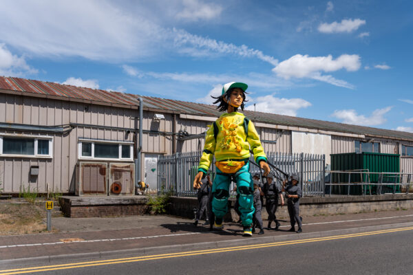 6m puppet, Zee, wearing a yellow tracksuit top, blue trackies and a blue hat; walking through an industrial estate. Three puppeteers helping her walk.