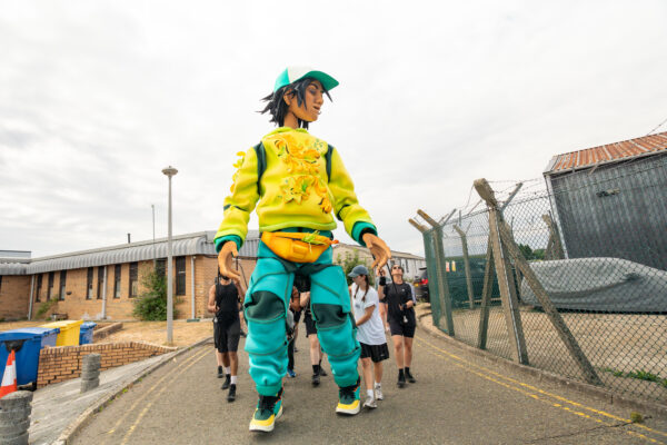 6m tall puppet, Zee, wearing a yellow tracksuit top and blue trackies. She is walking through an industrial park with three people guiding her.
