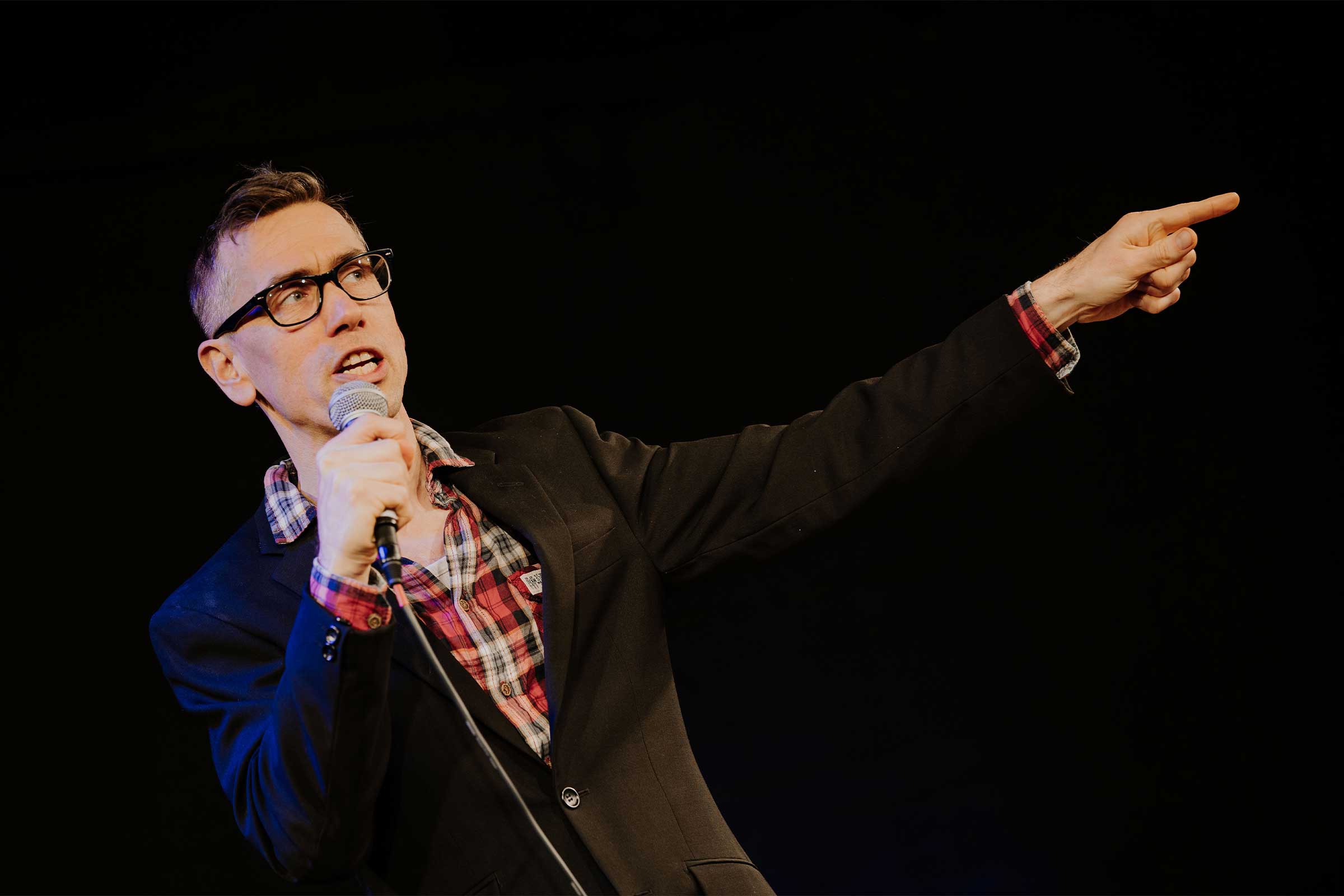 A mani in a checked shirt, suit jacket and glasses talks into a microphone whilst pointing with his arm outstretched