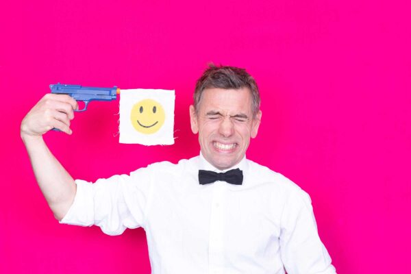 man wearing a bow tie, holding a gun to his head with a yellow smiley face shooting from the gun