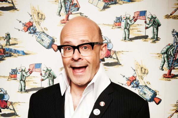 Comedian Harry Hill smiles against a patterned background. He wears a big smile.