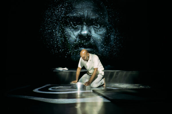 Man on his knees with a metal bowl infront of him, there is a light of a face behind him