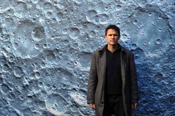 A photograph of a smartly dressed man stood in front of an image of the surface of the moon