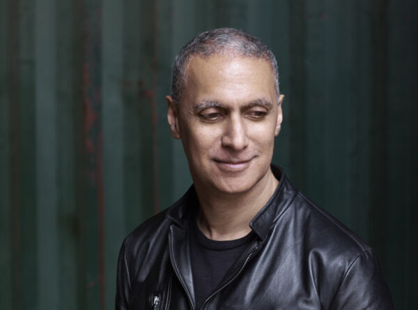 Man wearing leather jacket, looking down and smiling in-front of a dark background.