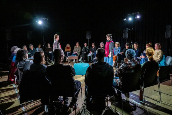 Two women stand on either side of the stage looking at each other with a blue suitcase between then. The audience surrounds them.