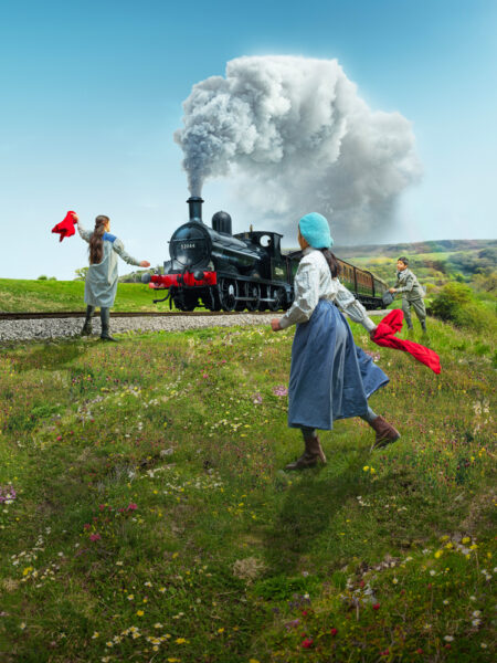 Three children in Victorian dress wave red flags at a steam train, on a sunny day in West Yorkshire.