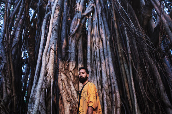 Male in yellow tunic, standing in-front of a large tree roots.