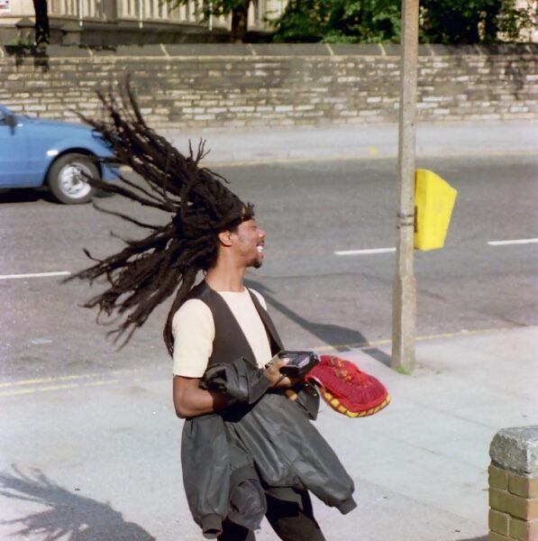 A man with long dreads flicks his hair, while standing on the pavement