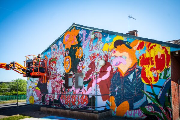 Vibrant mural on a wall, featuring colourful flowers, Billy Bantam and a Fox eating sausages