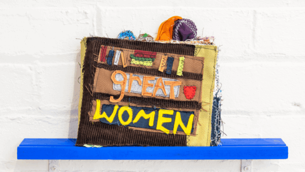 A textile art piece of a bookshelf with words 'great women' stitched over.