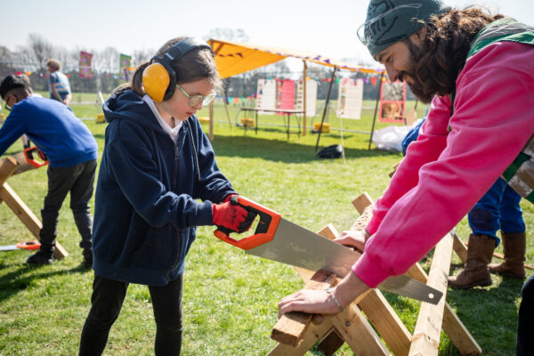 Child with ear defenders on sawing a piece of wood while an adult holds it.