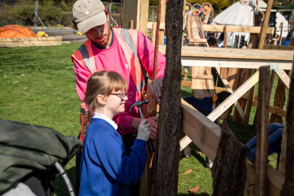 Young girl hammering a nail into a piece of wood. Adult supervisor in a pink high-vis helping her do it.