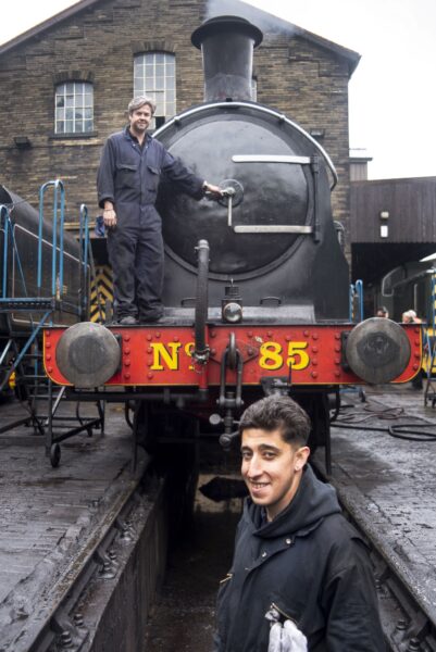 man stood infront and on a steam train and a younger man stood below and infront of the train smiling on the tracks