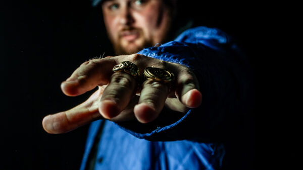 Male holding his hand in front of the camera. Wearing a lot of rings and a blue sports jacket.
