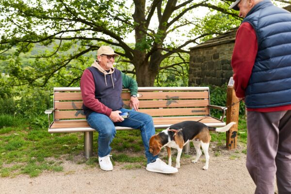 two men chatting, one has a dog, the other is sat on a bench