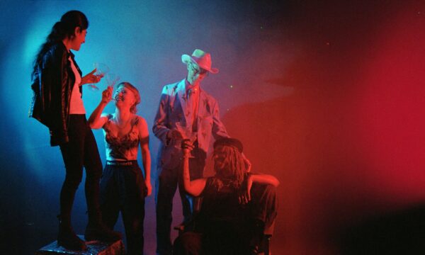 A film photograph - lit in blue and red shows four people toasting the camera with clear glasses.