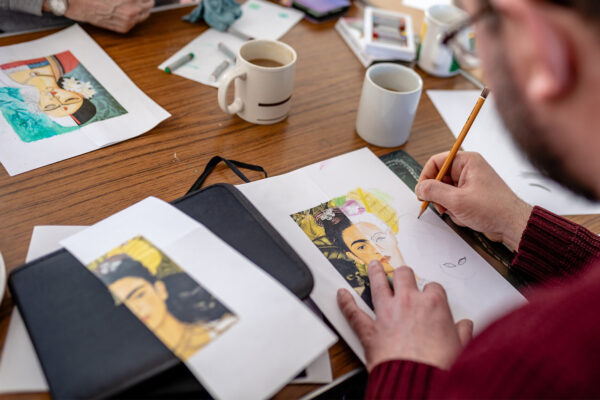 Man drawing on a table, table is filled with pencils, coffee