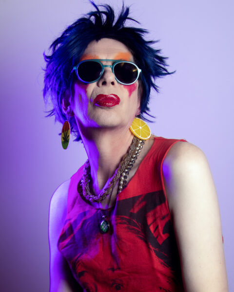 A person poses in front of a purple tinted wall, they are wearing bright make up beneath a pair of sunglasses and a red top.