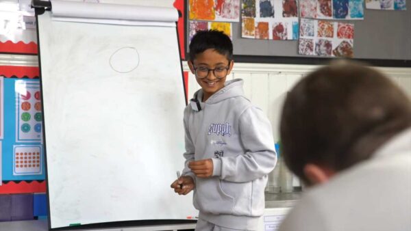 A young boy stands in a classroom drawing on a whiteboard. Is smiling and looking towards an adult who is copying his drawing.