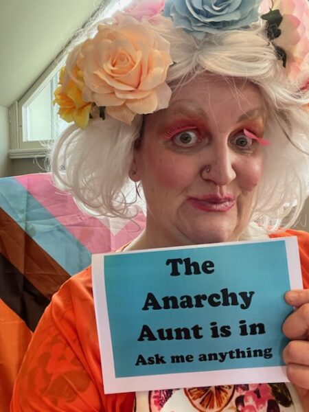 A person poses in a white wig with flowers, holding a sign that reads 'The Anarchy Aunt is in - ask me anything.'