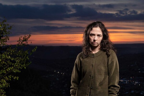 A moody woman in a green hoodie stands in front of a sunset landscape
