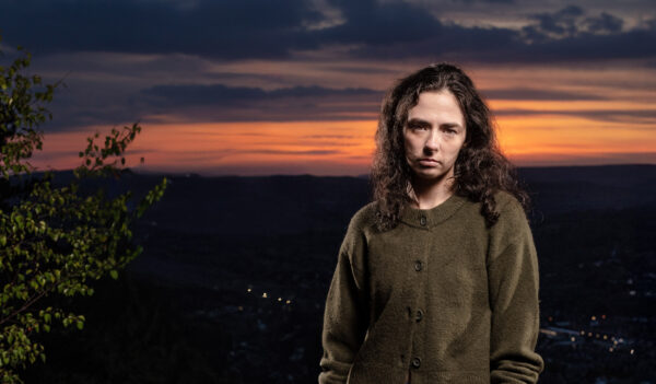 A moody woman in a green hoodie stands in front of a sunset landscape