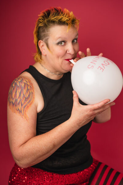 A person with a yellow and orange mullet poses in front of of a red wall, inflating a white balloon.